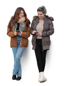 two women leaning against the wall and looking at their phones